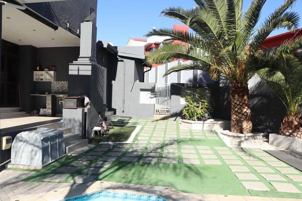 A well-maintained rear garden area featuring a paved patio, artificial lawn, mature palm tree, and a built-in outdoor braai area.