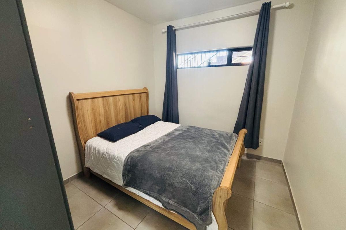 A compact bedroom featuring neutral tiled flooring, plain walls, and a high-set horizontal window with security bars.