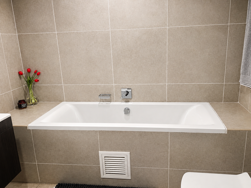 A modern bathroom featuring a built-in bathtub with neutral wall tiling, chrome fixtures, and a ventilation grille.