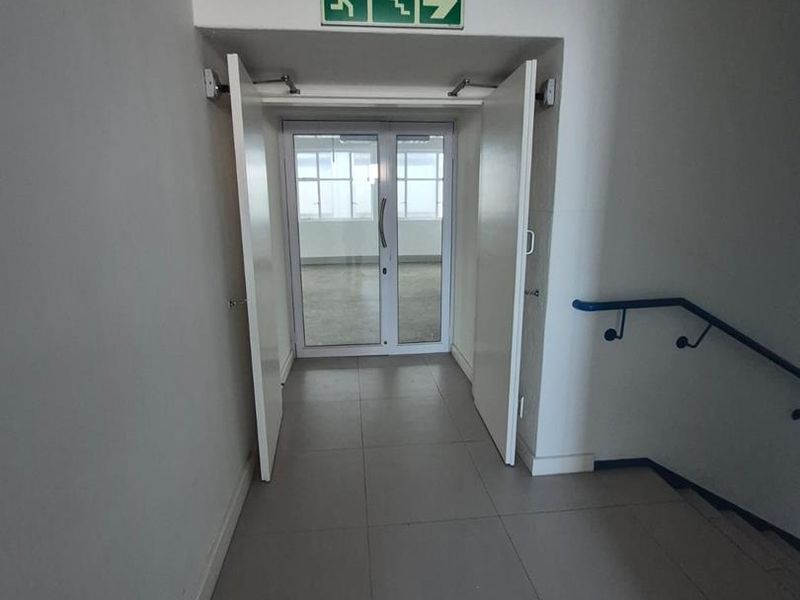 A functional office hallway featuring tiled flooring, a staircase handrail, and clear emergency exit signage leading to a glass-panelled doorway.