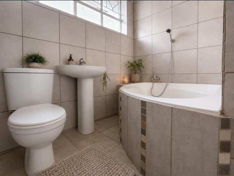 A well-maintained bathroom featuring a pedestal basin, toilet, and a corner bathtub with tiled finishes and natural light from a high window.