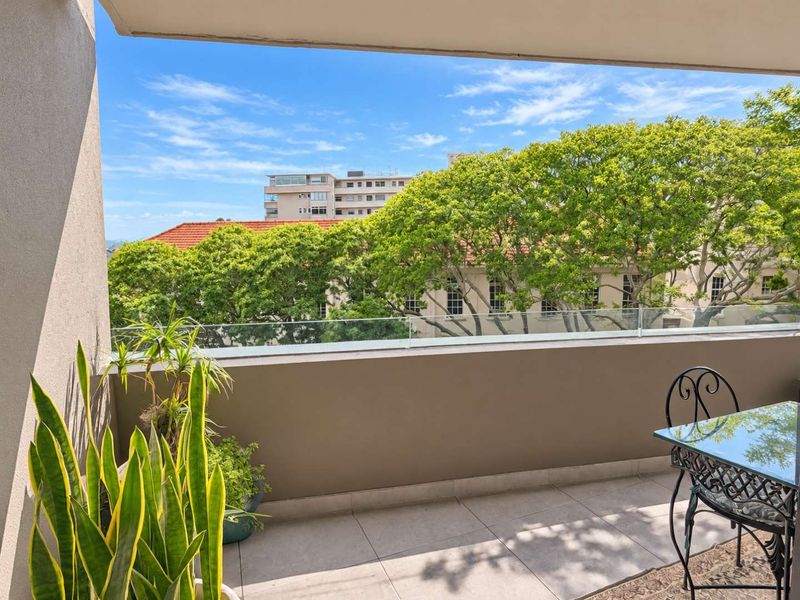 A well-maintained balcony featuring a modern glass balustrade and tiled flooring, offering an open view of the surrounding neighborhood and mature trees.