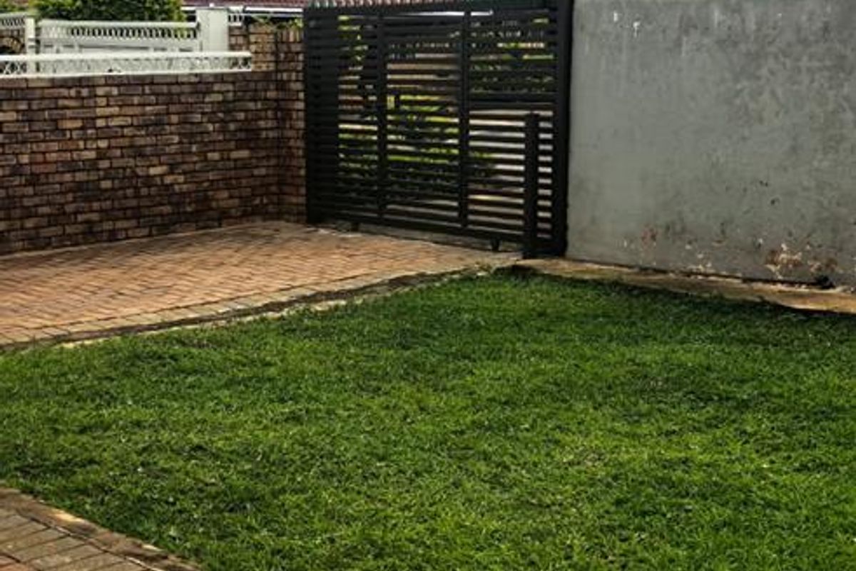 A private rear garden featuring a paved patio area, a small lawn, and a secure metal gate set within brick and plastered boundary walls.