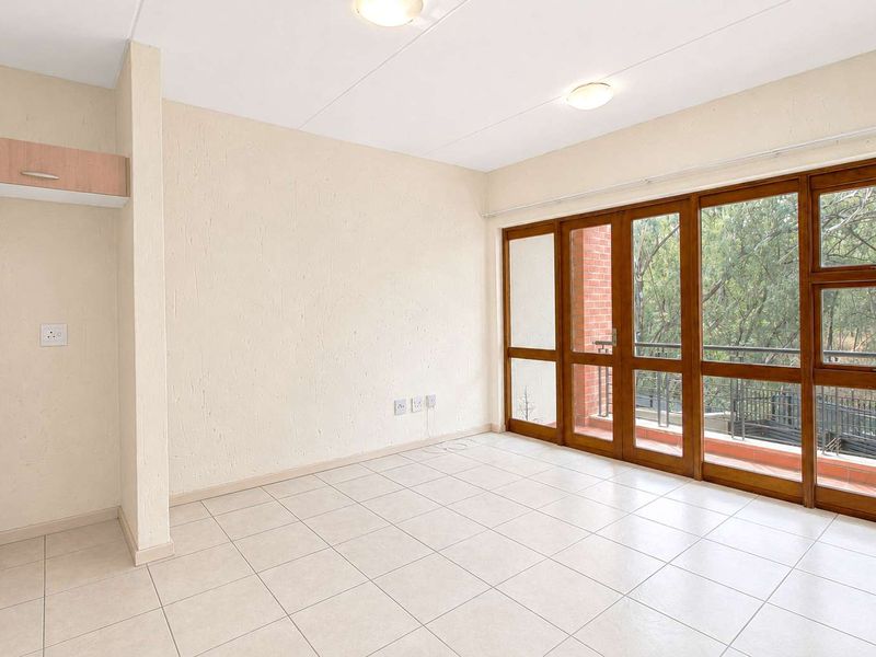 A bright, empty living area featuring neutral tiled flooring and large timber-framed glass doors opening onto a balcony with leafy views.