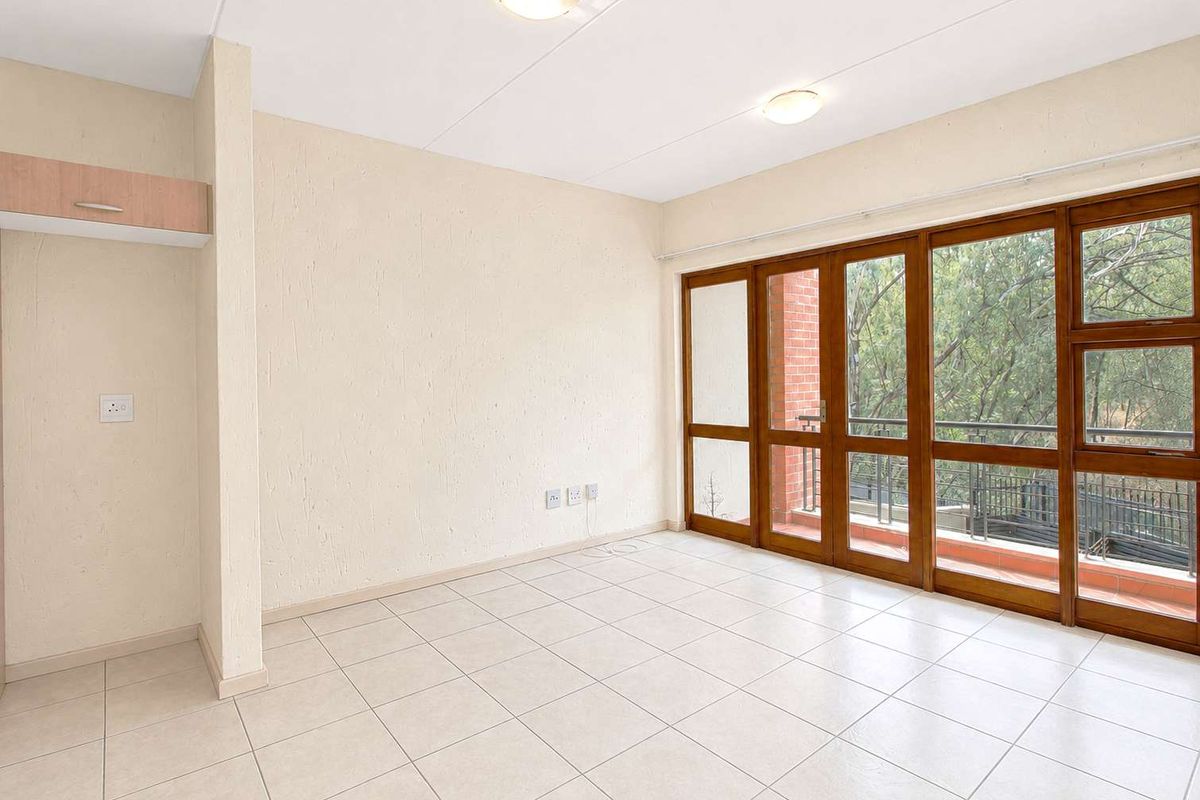 A bright, empty living area featuring neutral tiled flooring and large timber-framed glass doors opening onto a balcony with leafy views.