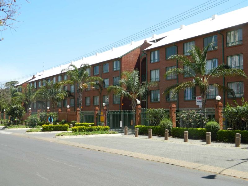 A multi-story brick apartment complex featuring a secure gated entrance, paved driveway, and established garden landscaping.