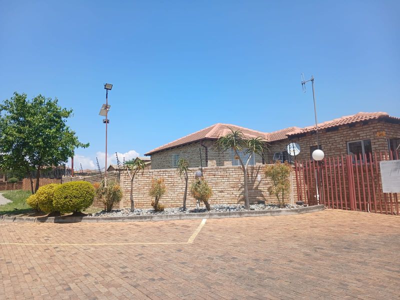 Front exterior view of a face brick apartment building featuring a paved driveway, secure boundary wall with electric fencing, and a metal entrance gate.