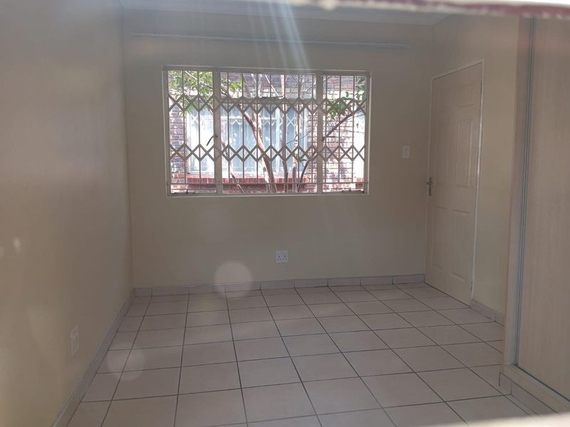 A clean, empty bedroom featuring neutral tiled flooring, a built-in cupboard, and a window fitted with a security gate.