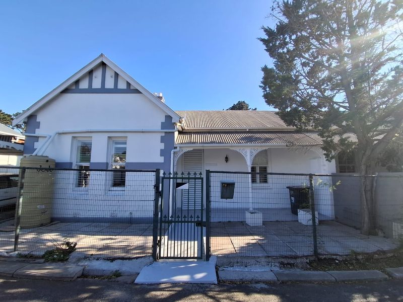 Front elevation of a single-story house featuring a covered veranda with decorative fretwork, a paved courtyard, and a corrugated metal roof.