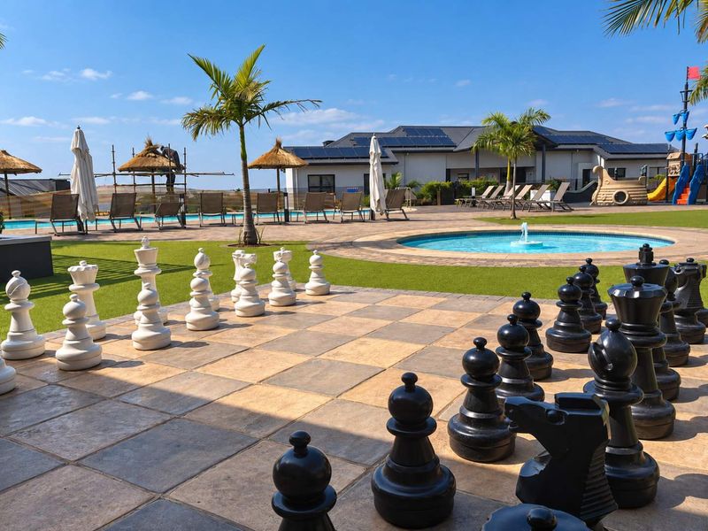 Outdoor recreational area featuring a swimming pool, water fountain, paved giant chess board, and landscaped grounds with solar-equipped building in the background.