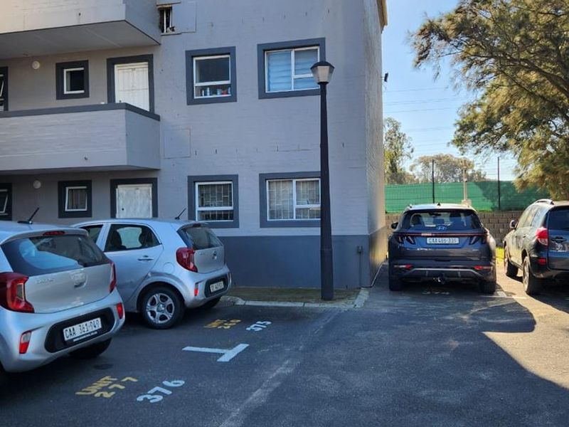 Exterior view of an apartment building facade featuring painted masonry, balconies, and an adjacent asphalt parking area with marked bays.