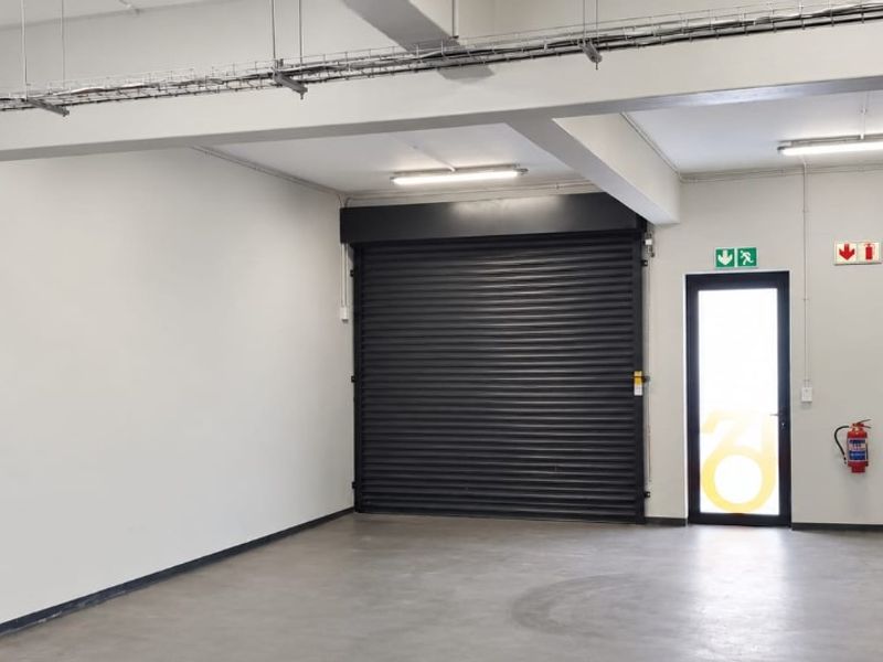 A clean, open-plan industrial utility space featuring a roller shutter door, exposed cable trays, and functional emergency safety equipment.