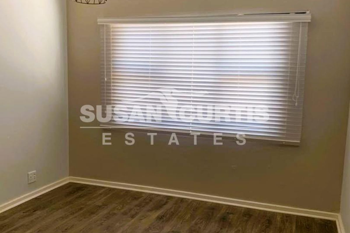 A clean, neutral bedroom featuring modern wood-look flooring, a wall-mounted air conditioning unit, and fitted venetian blinds.