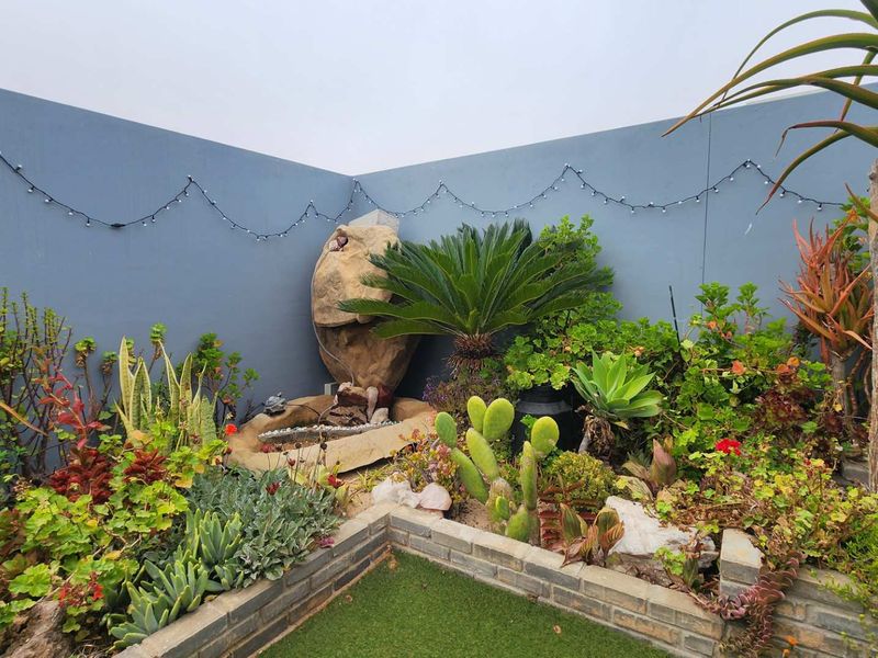A well-maintained garden corner featuring a stone water feature, established succulent landscaping, and a brick-edged artificial lawn against a painted boundary wall.