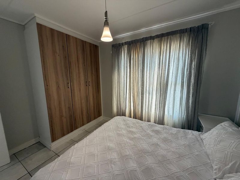 A well-maintained bedroom featuring built-in wooden cupboards, tiled flooring, and a neutral color palette with natural light filtering through window curtains.