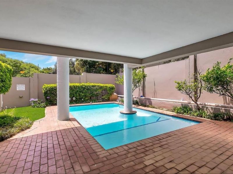 A private outdoor pool area featuring a covered patio with brick paving, structural columns, and a landscaped garden surround.