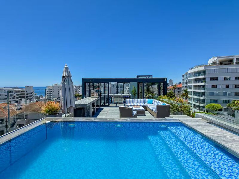 A luxury penthouse rooftop terrace featuring a swimming pool, built-in outdoor kitchen, and a glass-enclosed structure with panoramic urban and coastal views.