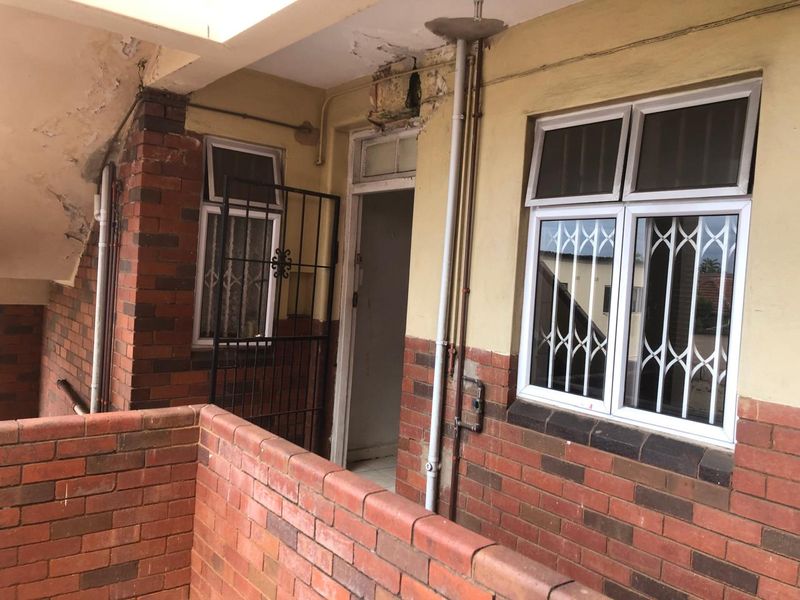 Exterior view of an apartment entrance featuring brickwork, security gates, and aluminium windows, showing signs of significant wear and maintenance requirements.