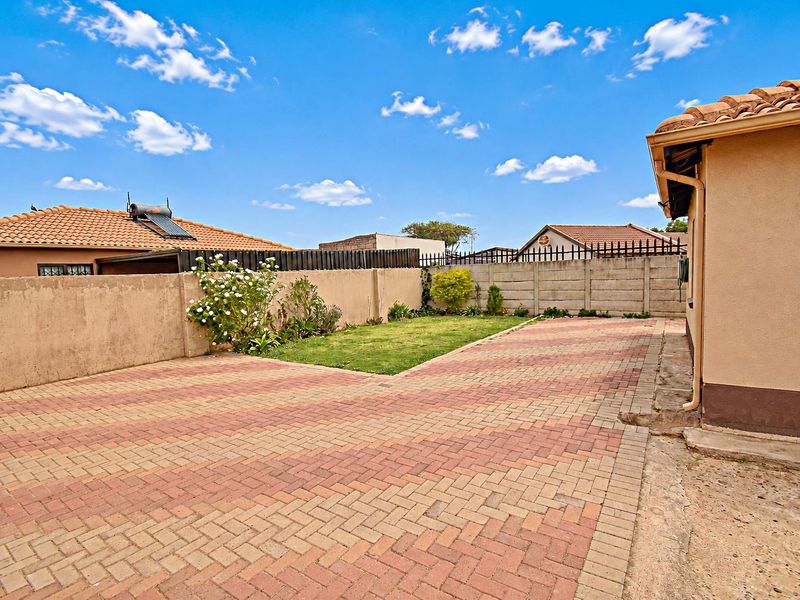 A rear exterior view featuring a paved driveway, a small lawn area, and a precast concrete boundary wall under a clear sky.