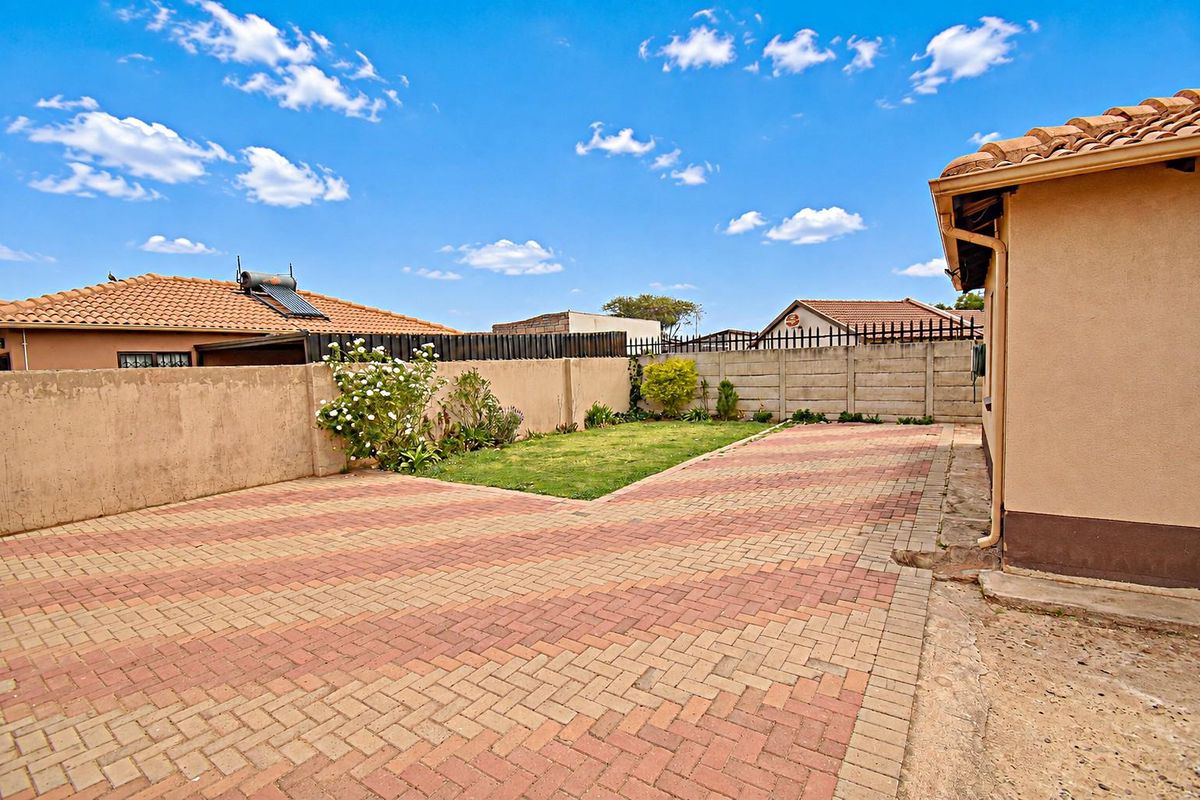 A rear exterior view featuring a paved driveway, a small lawn area, and a precast concrete boundary wall under a clear sky.