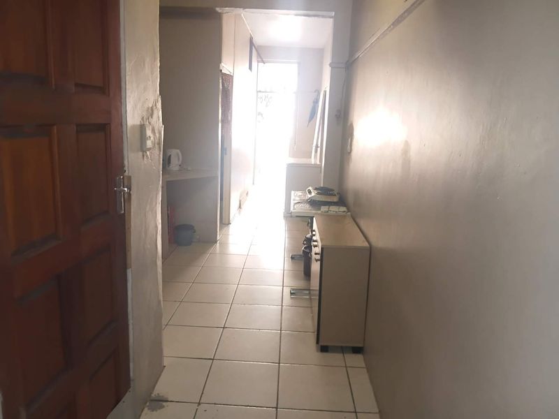 A narrow hallway featuring tiled flooring and a timber entrance door, leading towards a bright, naturally lit area at the end of the corridor.