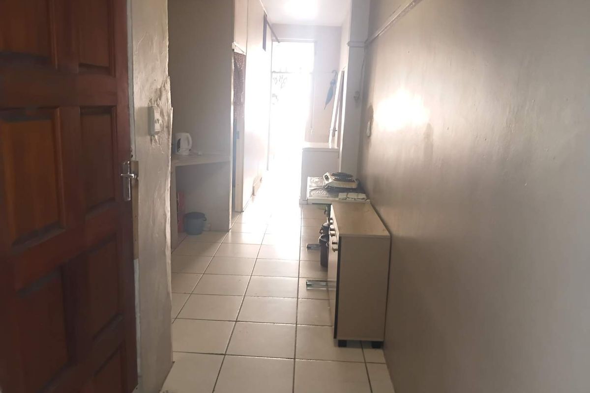 A narrow hallway featuring tiled flooring and a timber entrance door, leading towards a bright, naturally lit area at the end of the corridor.