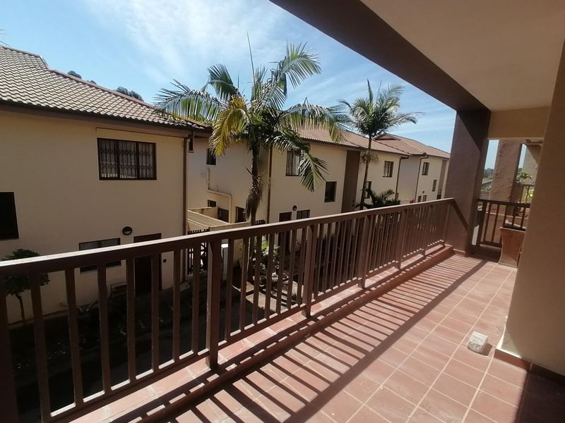 A covered balcony with tiled flooring and a railing, overlooking the townhouse complex grounds.