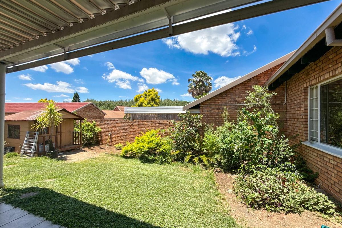 A private rear garden featuring a lawn, established shrubbery, brick boundary walls, and a covered patio area under a corrugated metal roof.