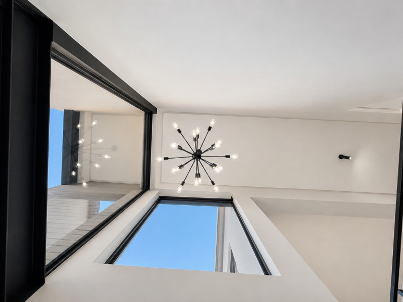 A bright, modern double-volume hallway featuring large windows, a contemporary chandelier, and a sleek glass balustrade.