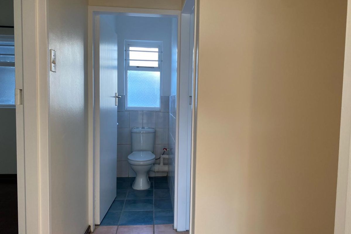 A small, functional bathroom featuring a toilet, tiled finishes, and a louvered window, currently presenting in a dated condition.