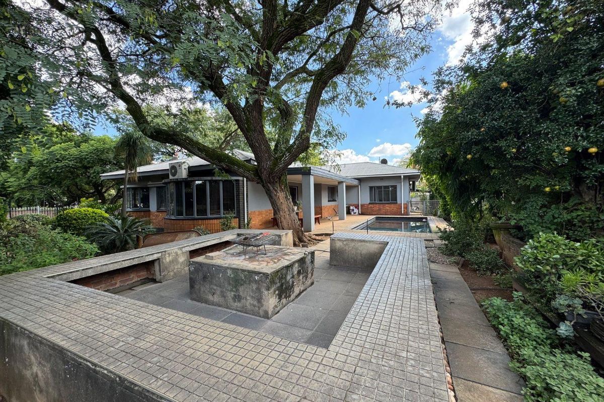 Spacious rear garden featuring a built-in fire pit area, paved patio, swimming pool, and mature trees, offering a private outdoor entertainment space.
