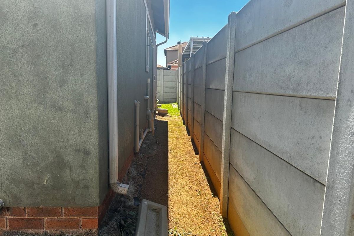 A narrow side passage between the house exterior and a precast concrete boundary wall, featuring exposed soil and external plumbing fixtures.