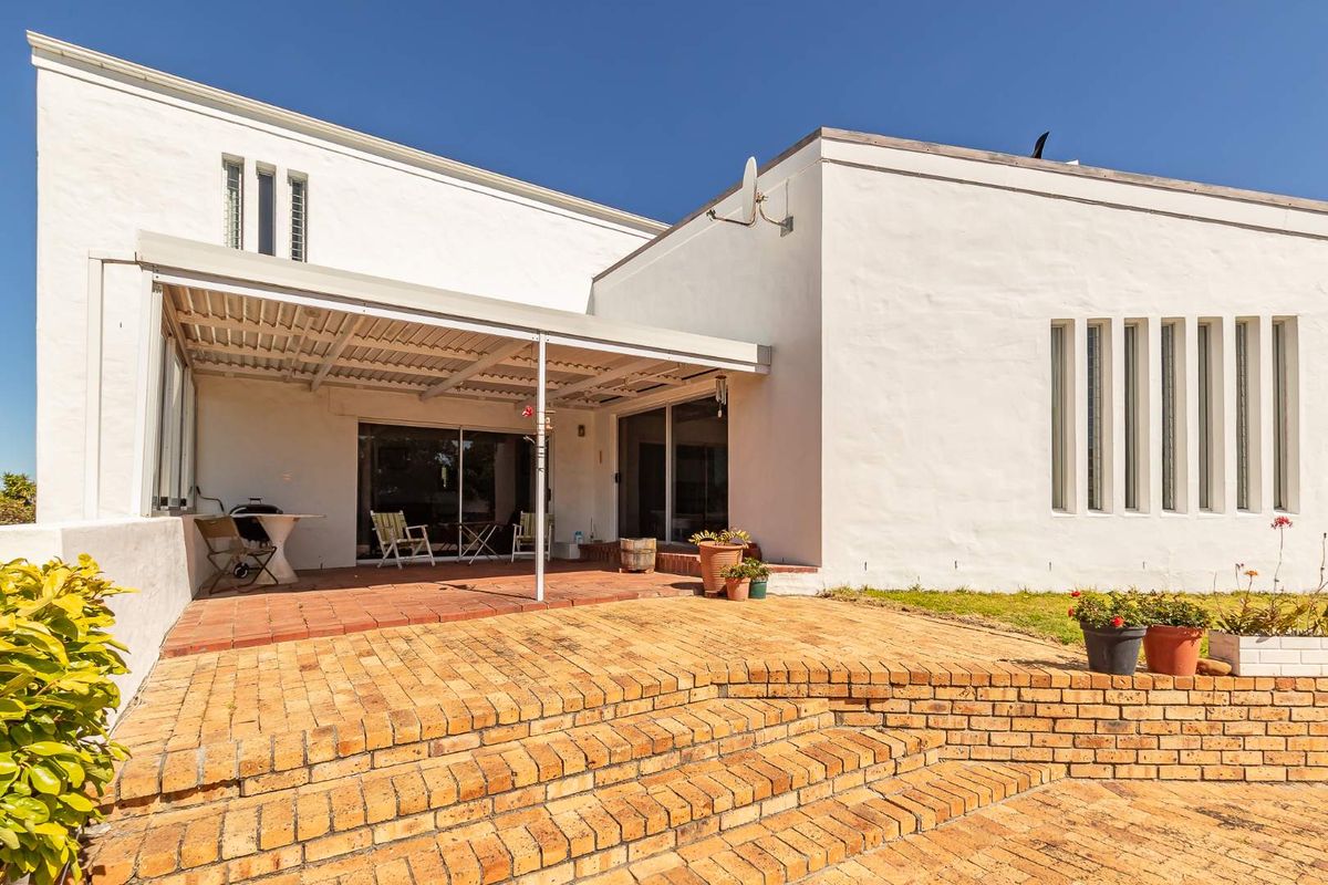 A rear exterior view featuring a covered patio area with brick paving, leading to a white-walled facade with vertical window detailing.