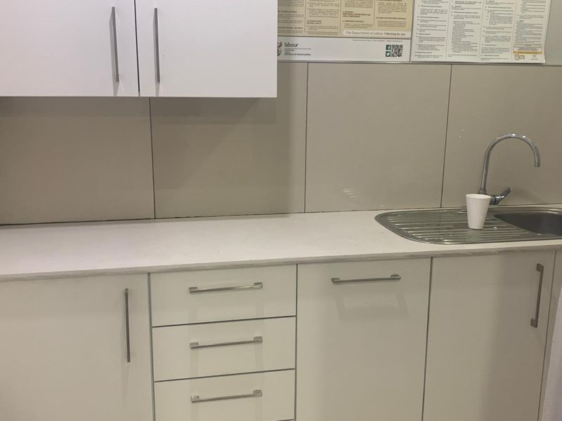 A clean, functional office kitchenette featuring white built-in cabinetry, a stainless steel sink with mixer tap, and a neutral tiled splashback.