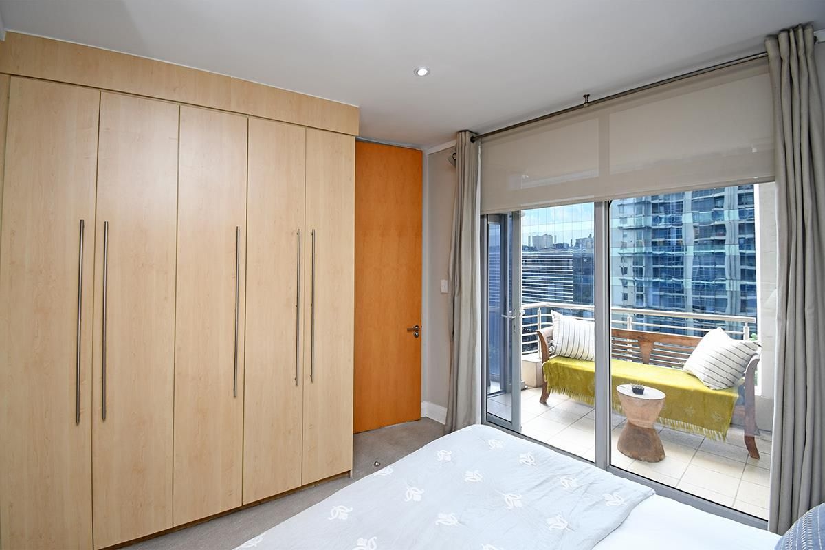 A bright bedroom featuring built-in light wood cabinetry, recessed ceiling lighting, and sliding glass doors opening onto a private balcony with city views.