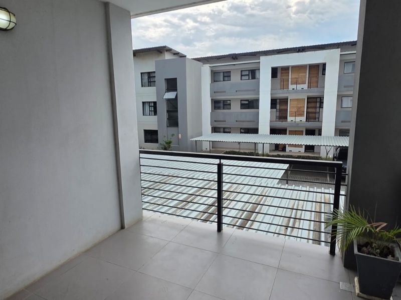 A covered balcony featuring tiled flooring and a metal balustrade, offering an open view of the apartment complex courtyard and parking area.