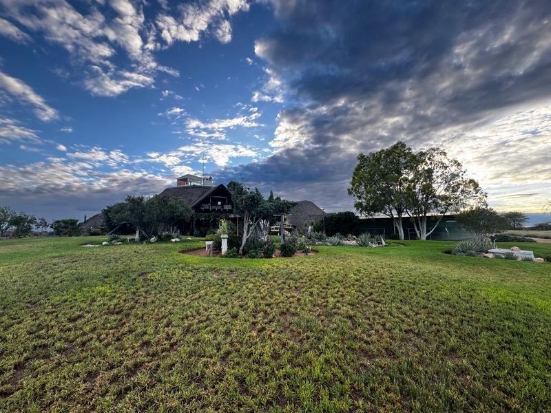 A wide-angle view of the rear exterior of a farm property featuring a traditional thatched roof and expansive, well-maintained garden grounds.