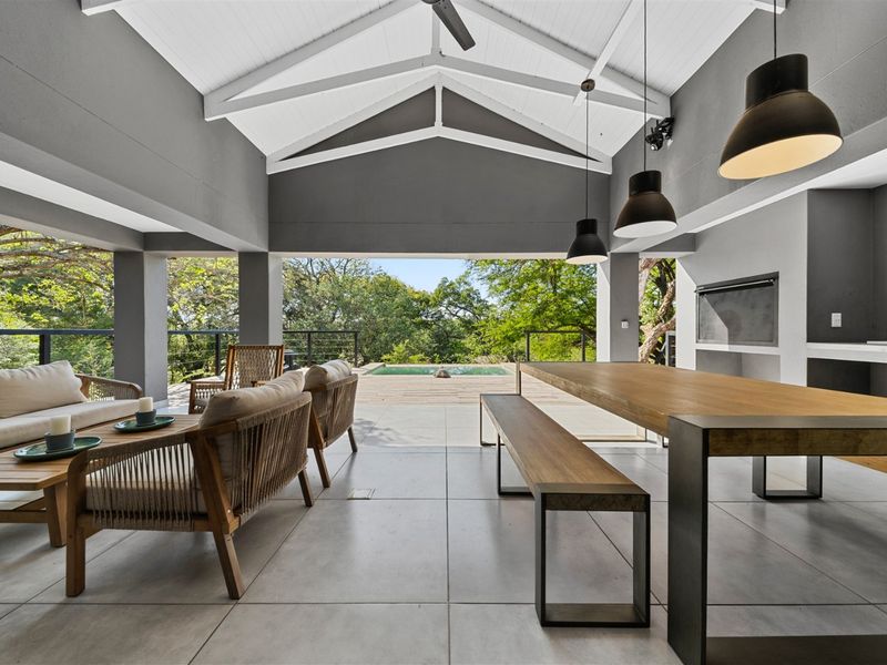 Spacious covered patio featuring a vaulted ceiling, built-in braai, and modern tiled flooring, seamlessly flowing out to the pool area and garden.