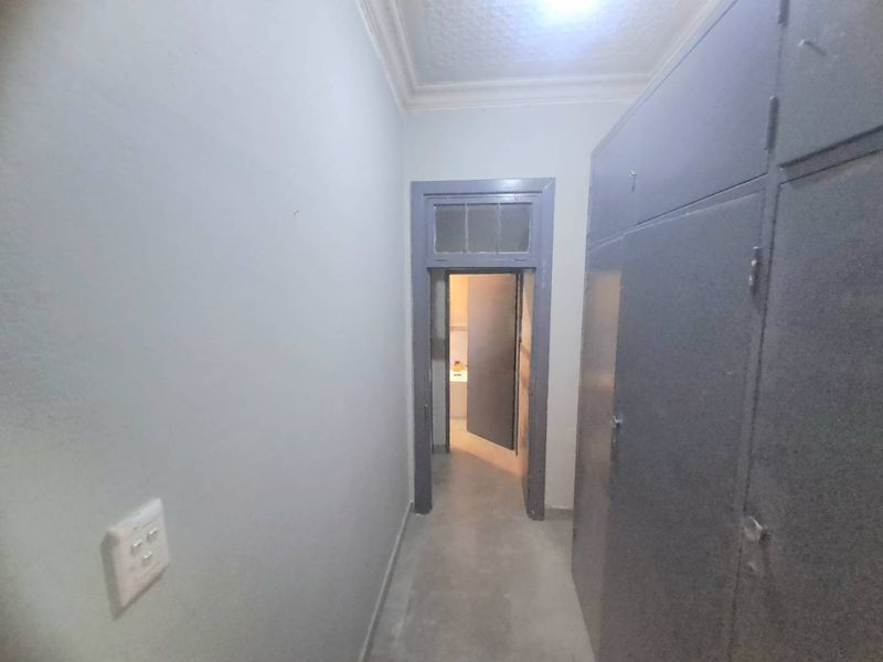 A narrow hallway featuring built-in storage cupboards, decorative ceiling cornice, and a transom window above the doorway leading to an adjacent room.