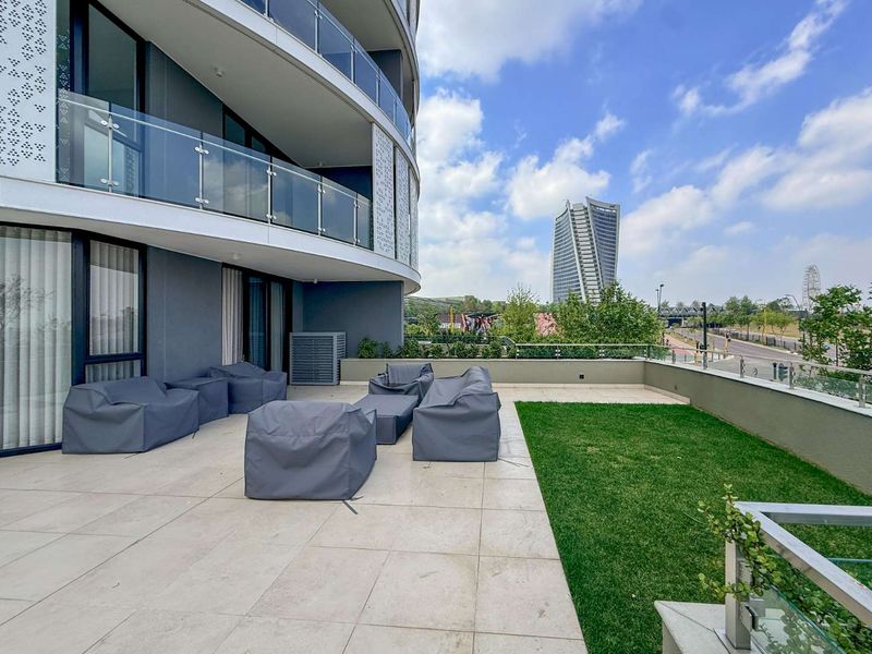 Spacious modern balcony featuring tiled flooring, a private lawn area, and glass balustrades with views of the surrounding urban landscape.