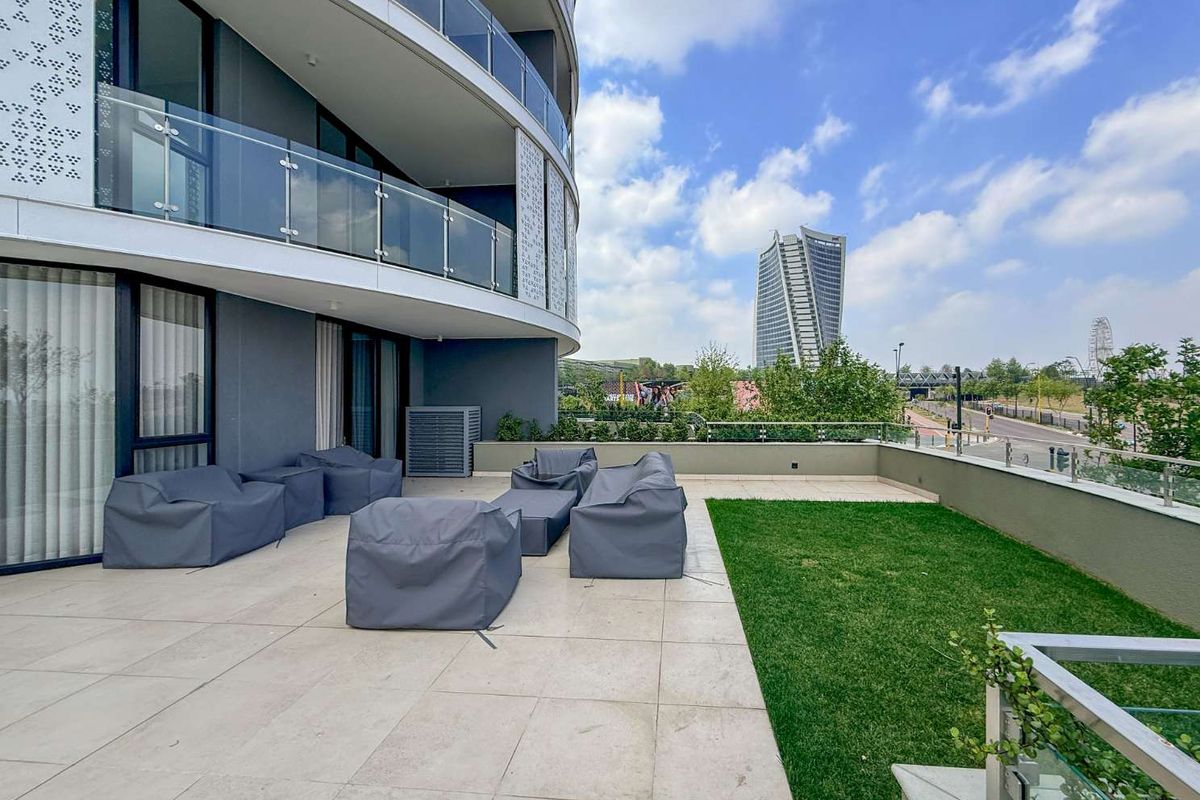 Spacious modern balcony featuring tiled flooring, a private lawn area, and glass balustrades with views of the surrounding urban landscape.