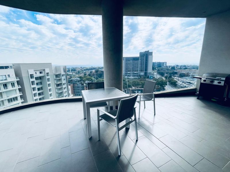 A spacious covered balcony featuring modern tiled flooring, a sleek glass balustrade, and expansive panoramic city views.