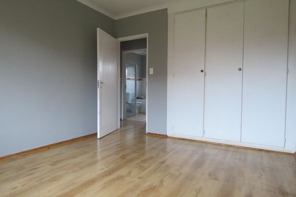 A bright, empty bedroom featuring light wood-look laminate flooring, built-in cupboards, and neutral grey walls, with an adjoining bathroom visible through the doorway.