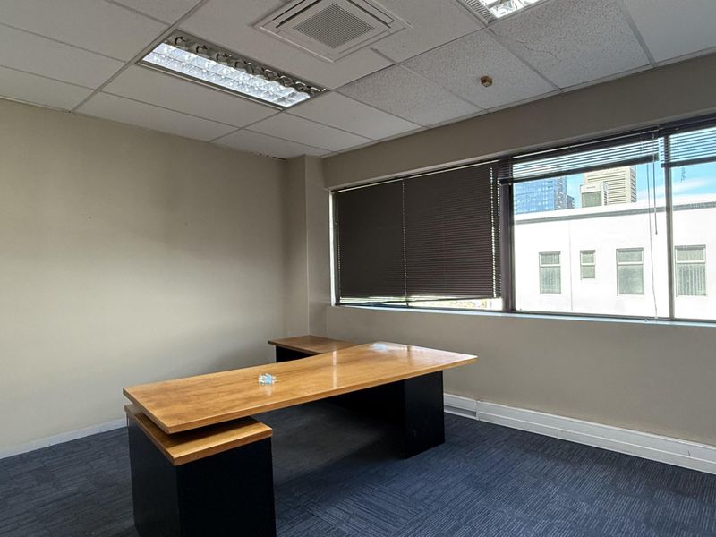 A functional office space featuring carpet tiles, a suspended ceiling with recessed lighting, and large windows with venetian blinds providing natural light.