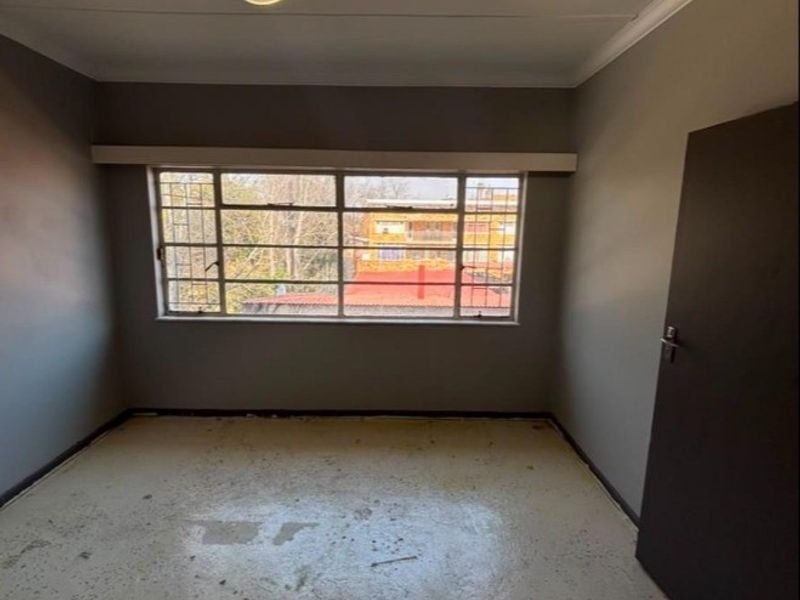 An empty bedroom featuring grey walls, a steel-framed window with security bars, and worn flooring, offering a blank canvas for renovation.