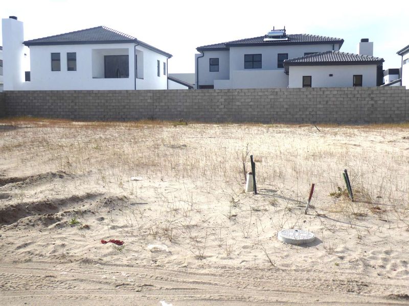 A vacant residential plot featuring sandy ground, utility service connections, and a concrete block boundary wall with neighboring houses in the background.