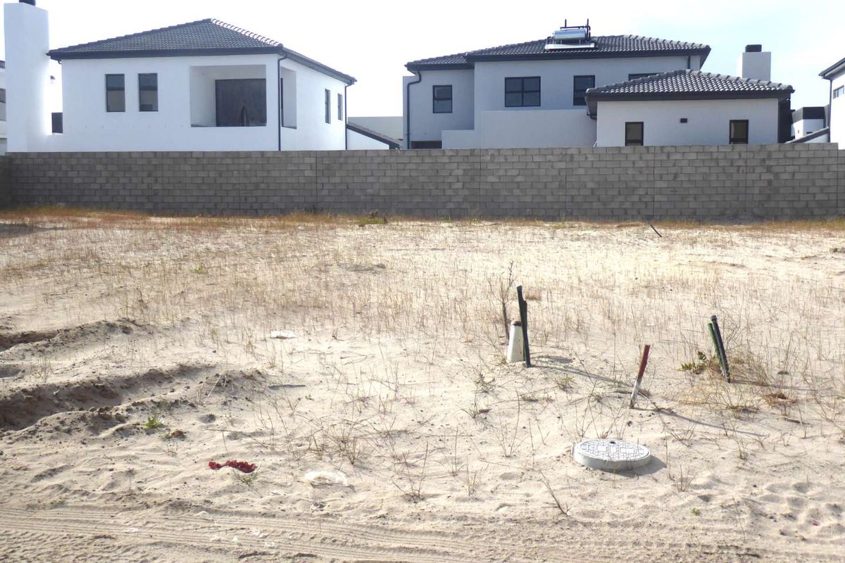 A vacant residential plot featuring sandy ground, utility service connections, and a concrete block boundary wall with neighboring houses in the background.