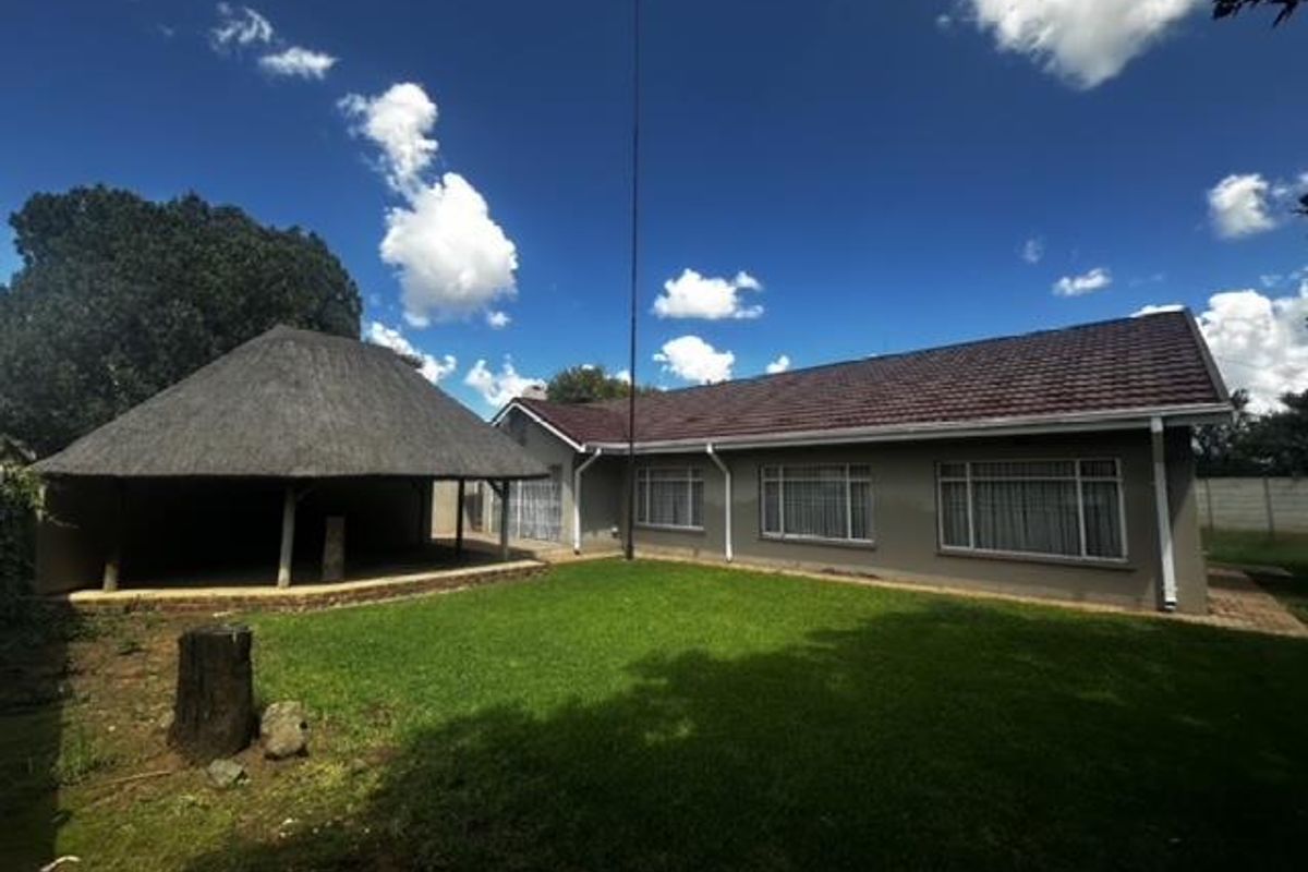 Rear view of the property featuring a spacious lawn, a traditional thatched lapa entertainment area, and a tiled roof house with large windows.