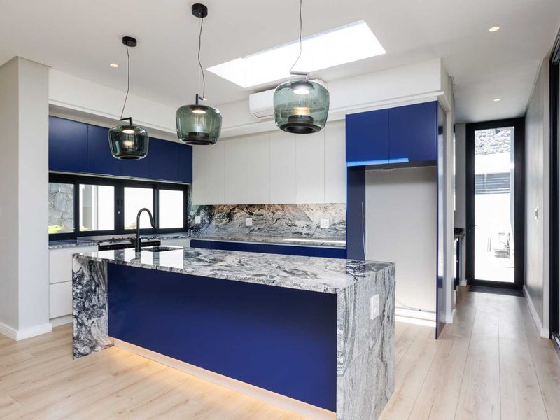 Modern kitchen featuring a striking blue and white cabinetry design, stone waterfall island bench, stone splashback, and a skylight for natural illumination.