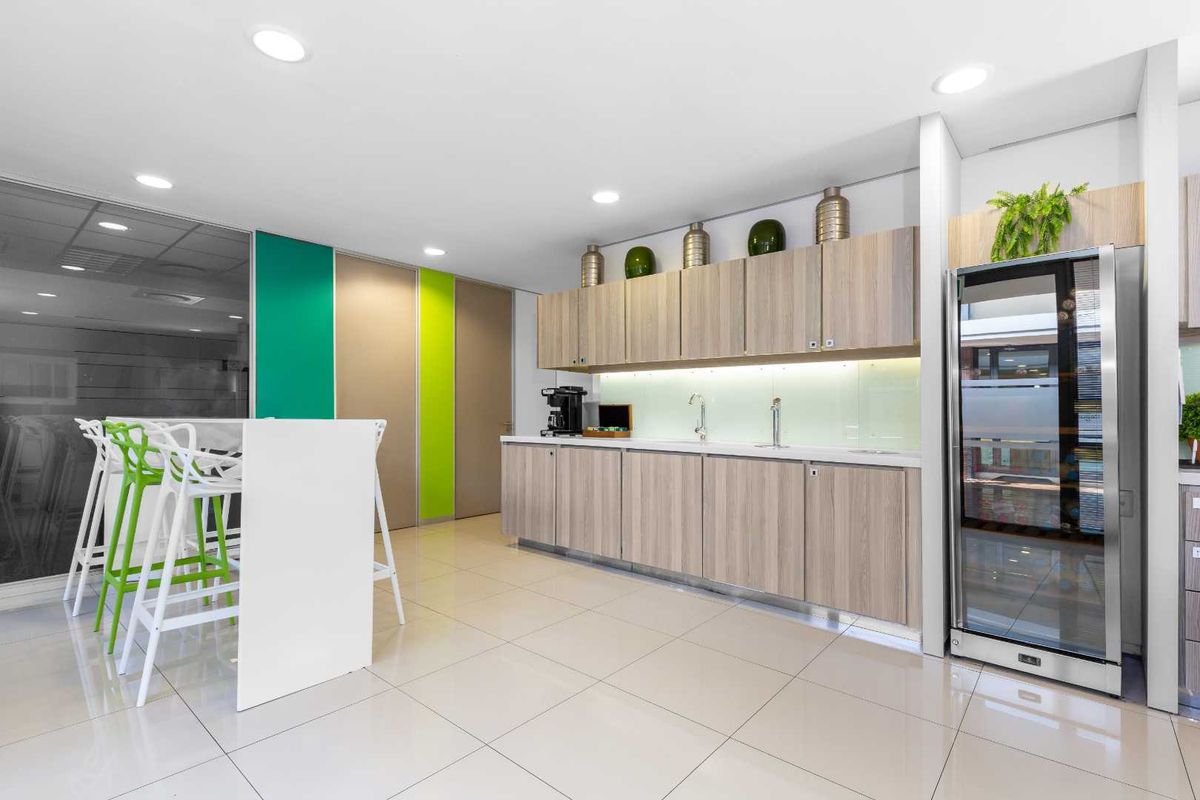 Modern office kitchenette featuring light wood cabinetry, integrated glass-door beverage cooler, sleek countertops, and recessed ceiling lighting.
